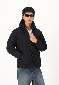 Chaqueta acolchada negra con capucha, con cierre de cremallera, dos bolsillos frontales, puños elásticos y una textura suave. Combinada con jeans grises.