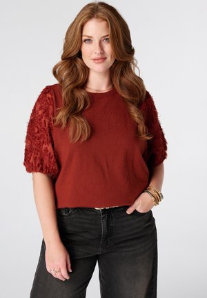 Vrouw met lang golvend rood haar, die een roestkleurige top met gestructureerde mouwen en een zwarte jeans draagt, glimlachend met één hand in de zak.