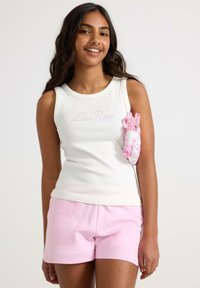 Hvid ribbet tanktop med "La Rose" i pink, parret med lyserøde shorts og et pink blomsteraccessory, der er stukket under armen.