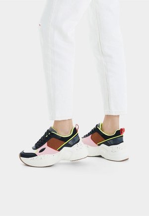 Jambes vêtues d’un pantalon blanc et de baskets épaisses noires, roses, marron et blanches avec des touches vert fluo et des languettes rouges au talon.