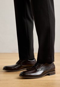 Zapatos de vestir de cuero marrón con un acabado suave, diseño con cordones y sutiles acentos laterales, combinados con pantalones negros a medida.