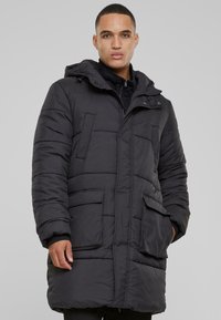 Crni zimski kaput s kapuljačom, sadrži četiri prednja džepa, zatvaranje na zip i kviltani dizajn za izolaciju i toplinu.