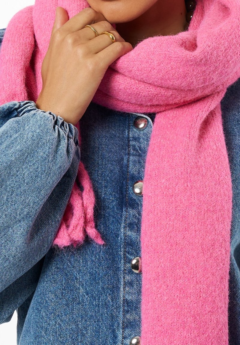 Femme portant une veste en denim et une écharpe en tricot rose vif, main près du visage avec des bagues en or et en argent visibles.