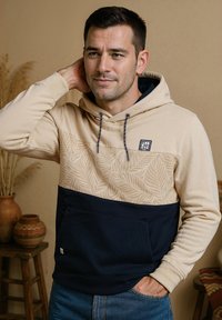 Hoodie met een beige bovenstuk met een bladerenpatroon, een donkerblauwe onderkant, een buidelzak en trekkoorddetails.