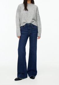 Suéter de punto gris con un cuerpo amplio y dobladillo acanalado, combinado con jeans oscuros de campana con cintura alta y puños deshilachados. Zapatos planos negros.