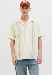 Camisa de manga corta, de color amarillo claro, hecha de una tela texturizada con rayas decorativas verticales en azul, rojo y amarillo. Cierre frontal con botones.