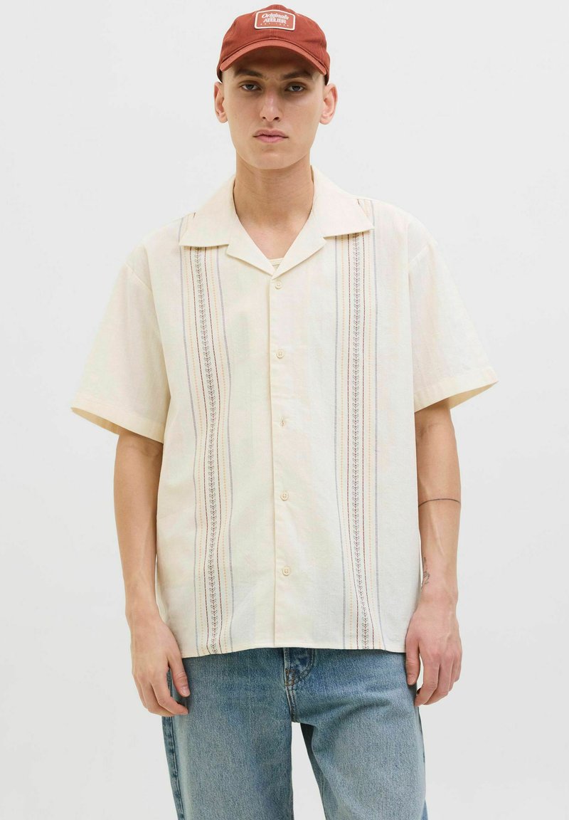 Camisa de manga corta, de color amarillo claro, hecha de una tela texturizada con rayas decorativas verticales en azul, rojo y amarillo. Cierre frontal con botones.