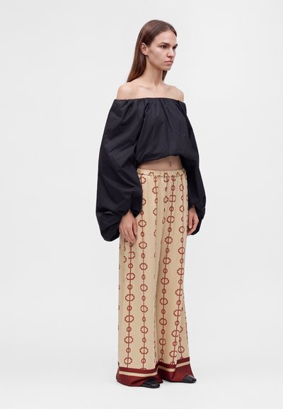 Femme portant un haut noir à épaules dénudées et manches bouffantes ainsi qu'un pantalon beige à jambes larges avec des motifs géométriques rouges et une bordure bordeaux.