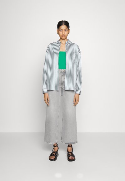 Camicia a righe con bottoni in grigio chiaro e blu navy, abbinata a una canotta verde, jeans larghi in grigio chiaro e sandali neri con cinturini.