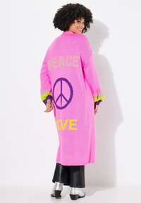 Langer rosa gestrickter Cardigan mit großem Text ��„PEACE“ und „LOVE“, einem Friedenssymbol und mehrfarbigen gestreiften Bündchen. Weiche Textur, oversized Passform.