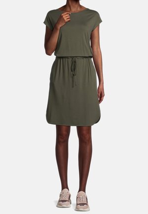 Frau trägt ein olivgrünes knielanges Kleid mit Kordelzug an der Taille und beigen Sportschuhen, steht vor einem schlichten hellen Hintergrund.