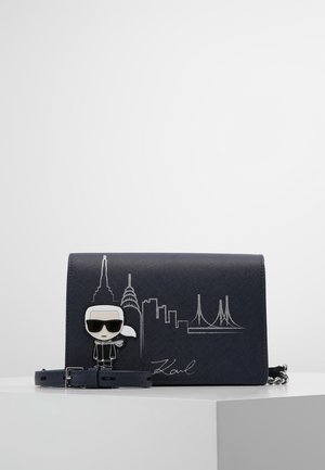 Marineblå crossbody taske med sølv bysilhuet broderi, tegneseriefigur med solbriller og justerbar rem på hvid overflade.
