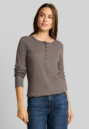 Junge Frau trägt ein braunes, gestreiftes Langarmshirt mit Knöpfen und blaue Jeans, steht vor einem grauen Hintergrund.