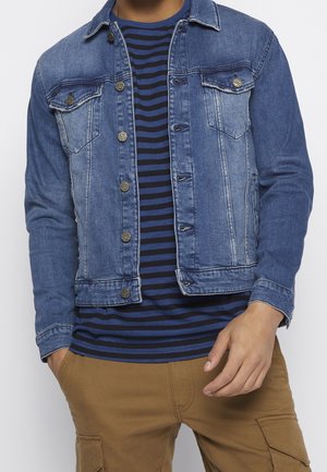 Homme portant une veste en denim bleu par-dessus une chemise rayée bleu marine et noire, avec un pantalon cargo beige, les mains détendues sur les côtés.