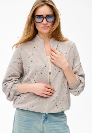 Woman with long blonde hair wearing large tinted sunglasses, a light beige knit zip-up sweater, and light blue jeans, posing against white background.