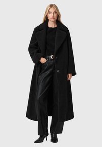 Černý, texturovaný dlouhý kabát s širokým klopem a velkými knoflíky, nošený přes černý top a kožené kalhoty, doplněný o boty na podpatku.