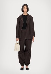 Un tailleur brun à rayures avec une coupe décontractée. La veste présente de grands boutons et un col cranté. Associé à un haut noir et à un sac de couleur claire.