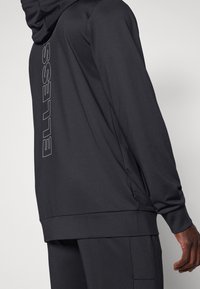 Homme portant un sweat à capuche noir à manches longues et un pantalon assorti, avec un audacieux logo vertical "ellesse" au dos du sweat.