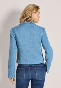 Lichtblauwe tweed jas met een getextureerde afwerking, getailleerde pasvorm en knoopaccenten op de mouwen, gecombineerd met donkere jeans, gezien vanaf de achterkant.