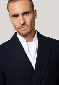 Granatowa wełniana marynarka z szpiczastymi klapami, przednią kieszenią i teksturowanym materiałem, nałożona na białą koszulę, charakteryzująca się schludnym, dopasowanym krojem.