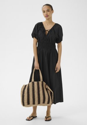 Vrouw in zwarte midi-jurk met pofmouwen die een grote geweven gestreepte tas vasthoudt, draagt zwarte teenslippers tegen een lichte achtergrond.