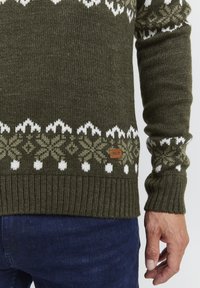 Pulover tricotat de culoare verde măsliniu cu modele de fulgi de nea albi și verzi deschiși, cu manșete și tiv din tricot cu nervuri, având un mic eticheta maro pe care scrie "ISOLID".