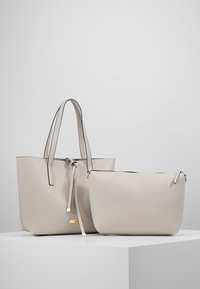 To lys grå håndtasker lavet af glat materiale. Den større taske har en snorelukning og lange håndtag, mens den mindre har en lynlåslukning.