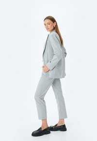 Helle graue maßgeschneiderte Anzugjacke mit einem Ein-Knopf-Blazer, schmal geschnittene Hose und schwarzen Slippern. Glattes Material mit subtiler Textur und klaren Linien.