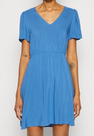 Femme portant une robe bleue à manches courtes, longueur genou, avec un décolleté en V et une taille froncée, montrant deux petits tatouages sur ses avant-bras.