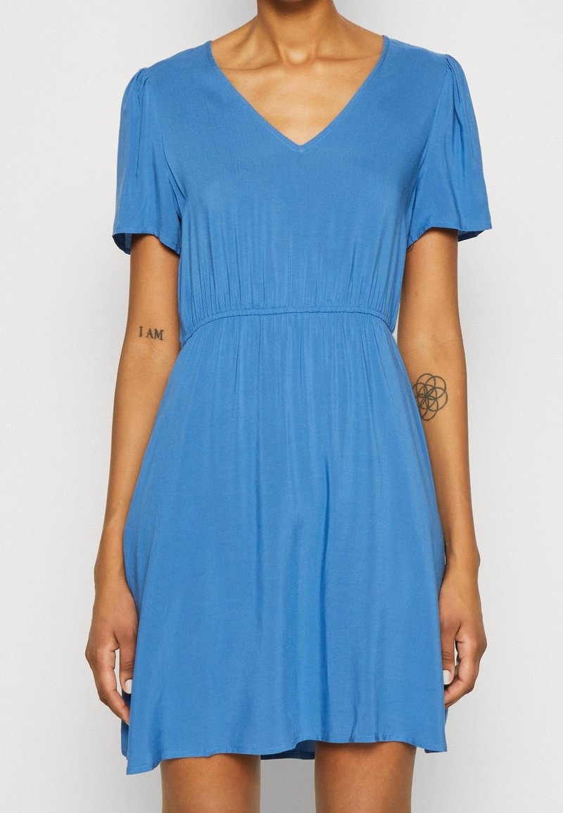 Femme portant une robe bleue à manches courtes, longueur genou, avec un décolleté en V et une taille froncée, montrant deux petits tatouages sur ses avant-bras.