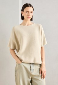 Lys beige strikkegenser med brede, korte ermer, avslappet passform og rund hals, kombinert med lyseblå denimjeans.