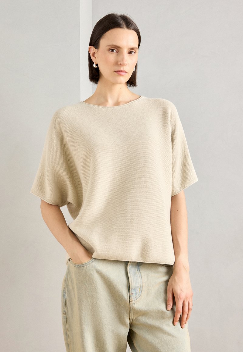 Lys beige strikkegenser med brede, korte ermer, avslappet passform og rund hals, kombinert med lyseblå denimjeans.