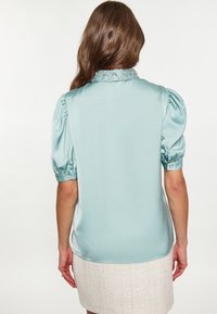Blusa di raso azzurro pallido con maniche a sbuffo e dettaglio del colletto con perline. Tessuto morbido, forma leggermente aderente e orlo curvato.
