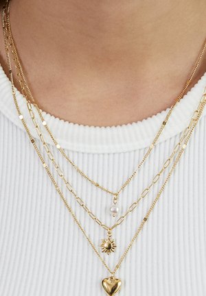 Four layered gold necklaces with varied chain styles; one has a pearl, one a sun charm, and one a heart pendant over a white ribbed top.