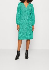 Grünes Wickelkleid mit schwarzen abstrakten Mustern, V-Ausschnitt, langen Ärmeln und einem Bindegürtel in der Taille, kombiniert mit schwarzen Knöchelstiefeln.