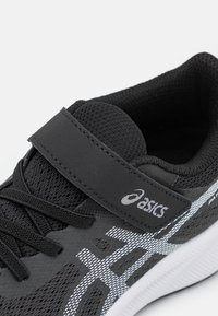 Baskets de sport noires en matériau en maille, avec lacets et sangle Velcro, présentant des accents et un logo blancs. Surface texturée et silhouette élégante.