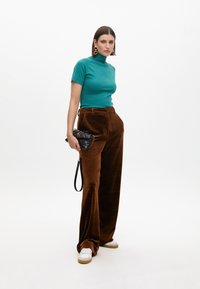 Maglione fitted color teal, pantaloni wide-leg in velluto marrone, sneakers bianche, borsa a tracolla nera trapuntata con dettaglio in catena dorata, modella in posa su sfondo bianco.