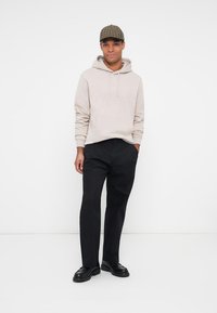 Hoodie beige chiaro con tasca frontale, abbinato a pantaloni neri e scarpe nere. Un berretto a quadri completa l'outfit casual.