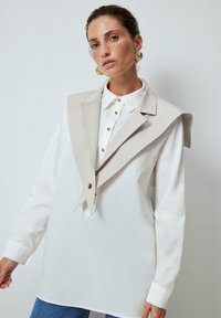 White shirt with fitted silhouette, featuring a beige oversized collar and gold button details on the front, worn with denim jeans.