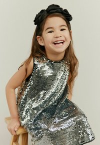 Strieborné flitrové šaty bez rukávov, s okrúhlym výstrihom a voľným siluetou. Čierny látkový čelenka s kvetinovými akcentmi.