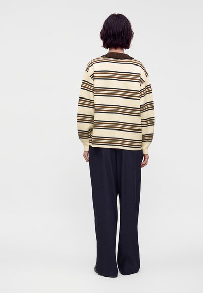 Personne debout, dos tourné, portant un pull crème avec un motif rayé noir et marron et un pantalon noir ample sur un fond clair uni.