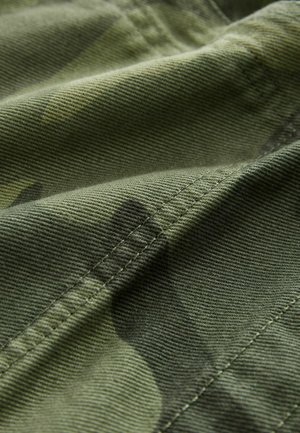 Green camouflage fabric with a textured finish, featuring subtle pattern variations and visible stitching along the edges.