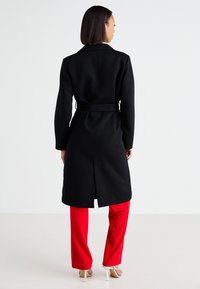 Black long coat with a belt, featuring a back slit. Worn over red pants, on a plain background. Smooth texture and tailored design.