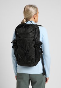 Zaino nero con più scomparti con zip, tasche laterali in rete, spallacci rinforzati e un design minimalista con una finitura testurizzata.