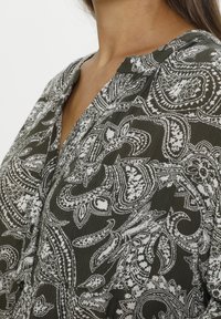 Chemise vert foncé à motif cachemire blanc, avec un col en V et des détails boutonnés. Le tissu est léger et texturé.