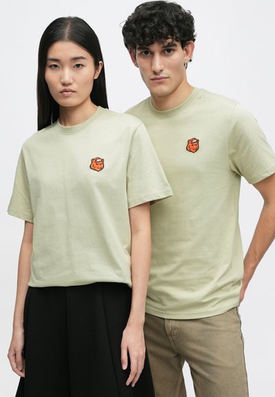 Ανοιχτό πράσινα βαμβακερά μπλουζάκια με στρογγυλή λαιμόκοψη και κοντά μανίκια, που διαθέτουν ένα πορτοκαλί κεντητό λογότυπο αρκούδας στο αριστερό στήθος.