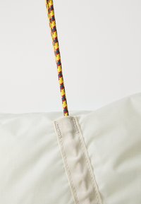 Lyst stoff med teksturert overflate, med en flettet, flerfarget tau festet på toppen. Forsterkede sømmer synlige.
