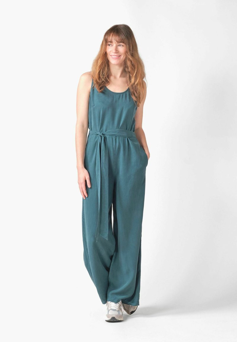 Femme aux longs cheveux portant une combinaison sans manches bleu sarcelle avec taille nouée et baskets blanches, debout et souriante, main droite dans la poche.