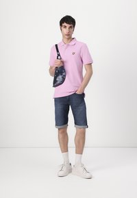 Rosa Polohemd mit einem kleinen gelben Logo, kombiniert mit hochgerollten Denim-Shorts und weißen Sneakern. Enthält eine marineblaue Batikhandtasche mit Träger.