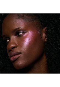 Nærbilde av en person med mørk hud og krøllete hår, som har glitrende rosa øyenskygge og highlighter på kinnbena.